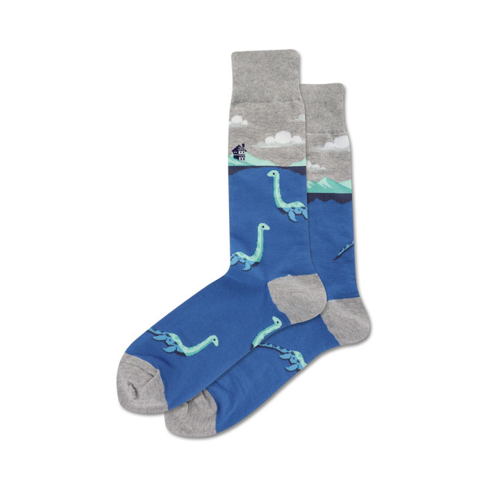 A pair of blue crew socks with a grey cuff. The socks feature a repeating pattern of light teal Loch Ness monsters swimming in the water, with a small house on a hill in the background. }}