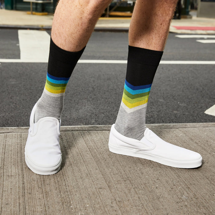 A man is crossing the street in front of a subway station. He is wearing a blue, green, and white striped shirt, white shorts, white sneakers, and colorful socks. He has a tattoo on his right arm.