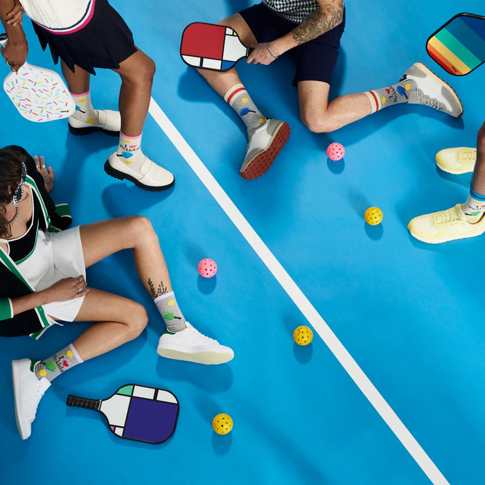 Overhead shot of several people sitting on a blue pickleball court wearing different colored socks, holding pickleball paddles and balls.