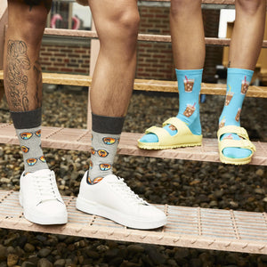 Two people are standing on a metal grate. The person on the left is wearing white sneakers, gray socks with a pattern of blue and pink sunglasses, and a tattoo of a flower on their leg. The person on the right is wearing yellow sandals, blue socks with a pattern of iced coffee drinks, and dark gray shorts.