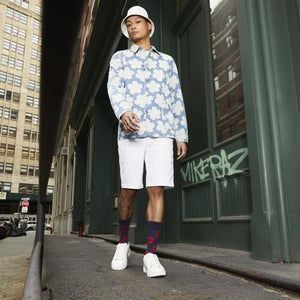 A young man is wearing a white bucket hat, a blue denim jacket with white flower pattern, white shorts, red socks, and white sneakers. He is standing on a city street with graffiti on the wall behind him.