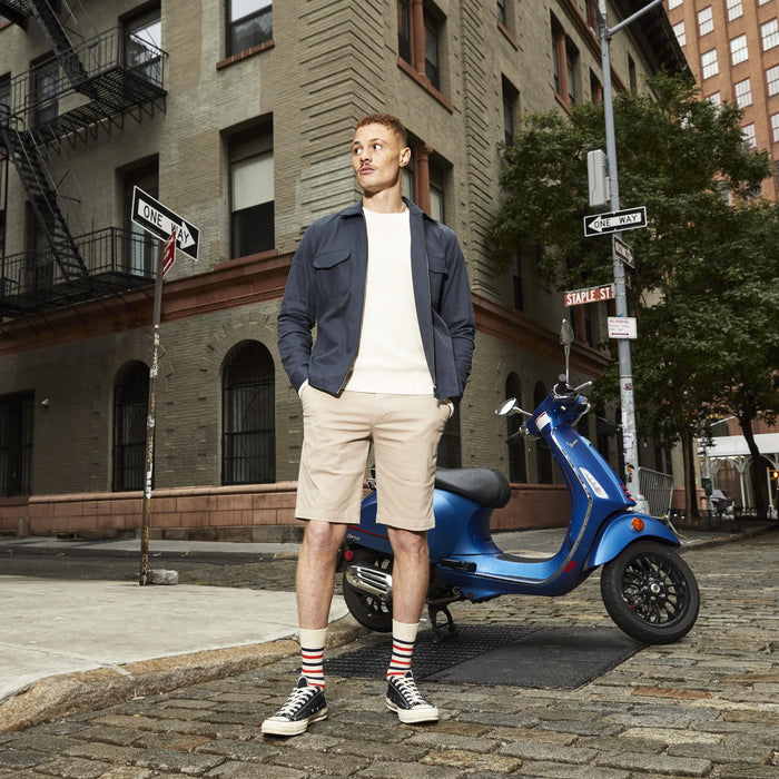 A man wearing a blue jacket, white sweater, khaki shorts, red and white striped socks, and black sneakers is standing in front of a blue Vespa scooter on a cobblestone street. There are buildings on either side of the street and a one-way sign in the background.