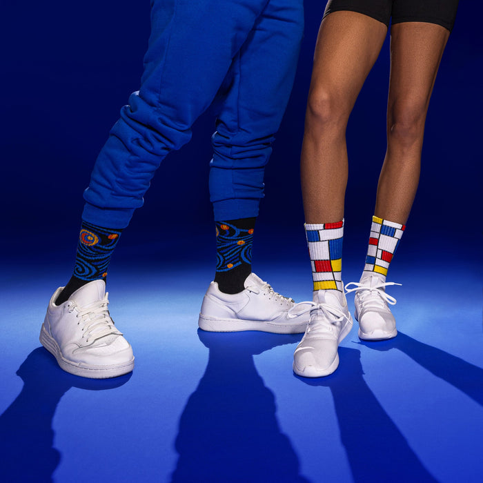 Lifestyle shot of two models wearing Starry Night and Mondrian-inspired socks with white sneakers against a blue background.
