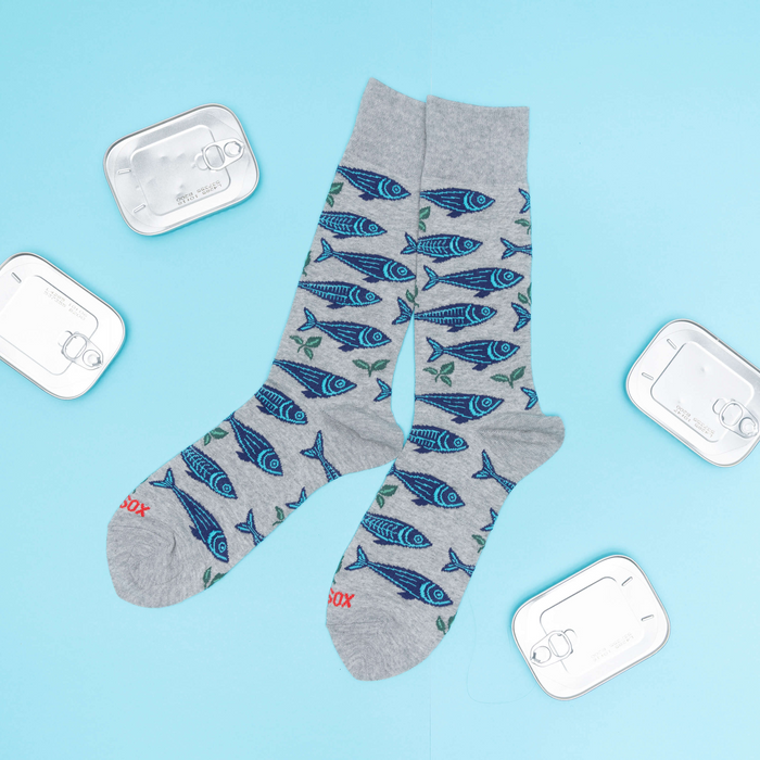 Grey sardine patterned crew socks laid flat on a blue surface, surrounded by four silver empty sardine cans.