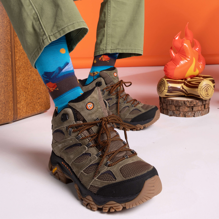 Person wearing green pants and hiking boots with scenic canoe and camping socks on a white floor.