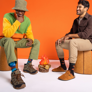 Two men sitting, one wearing 'Dam It' beaver socks with a brown shirt, next to a faux campfire against an orange wall.
