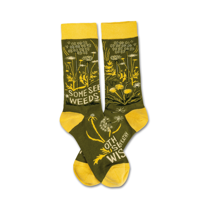 Pair of olive green crew socks with yellow accents. Features a dandelion and wildflower design and the text 'Some See Weeds'. }}