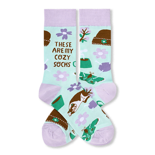 A pair of novelty crew socks with a pastel mint green background, lavender cuffs and toes, and a whimsical pattern of brown hats, purple flowers, green boots, butterflies, and a small dog. The text 'THESE ARE MY COZY SOCKS' is printed on one sock.