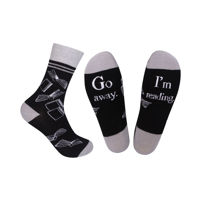 A pair of black crew socks with a grey cuff. The socks feature a white design of open books and the phrases 'Go Away' and 'I'm Reading'.