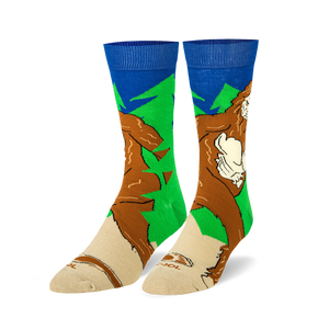 Pair of crew socks featuring a Sasquatch design.  Colors include brown, green, and blue. The Bigfoot image is walking through a forest.