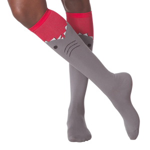 A pair of gray knee-high socks with a red cuff. The socks have a cartoon shark face on them, with the shark's mouth open and its teeth showing.