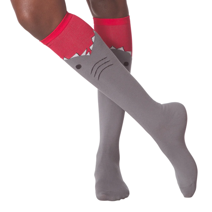 A pair of gray knee-high socks with a red cuff. The socks have a cartoon shark face on them, with the shark's mouth open and its teeth showing.