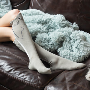 A person wearing gray knee-high socks with a shark design on them. The socks have a white tip at the toe and a white line that runs along the side of the leg. The person is sitting on a brown leather couch with a fluffy blue blanket with white spots on it.