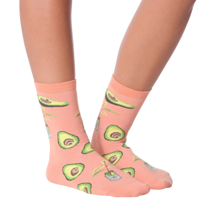 A person is sitting on a stool with their feet resting on the rung below. They are wearing light blue distressed jeans and pink socks with an avocado pattern. The socks are mid-calf length and the pattern features green avocados with brown stems and a tiny white flower.