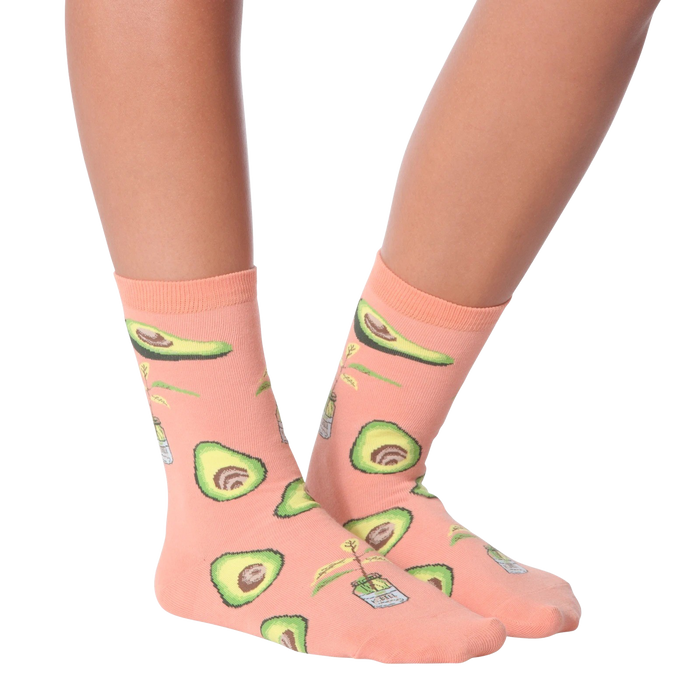 A person is sitting on a stool with their feet resting on the rung below. They are wearing light blue distressed jeans and pink socks with an avocado pattern. The socks are mid-calf length and the pattern features green avocados with brown stems and a tiny white flower.