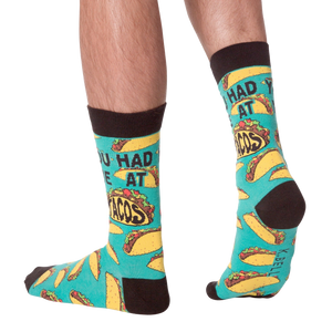Lifestyle photo showing the 'You Had Me at Tacos' socks on a person's feet. The teal and yellow sock pattern is visible.