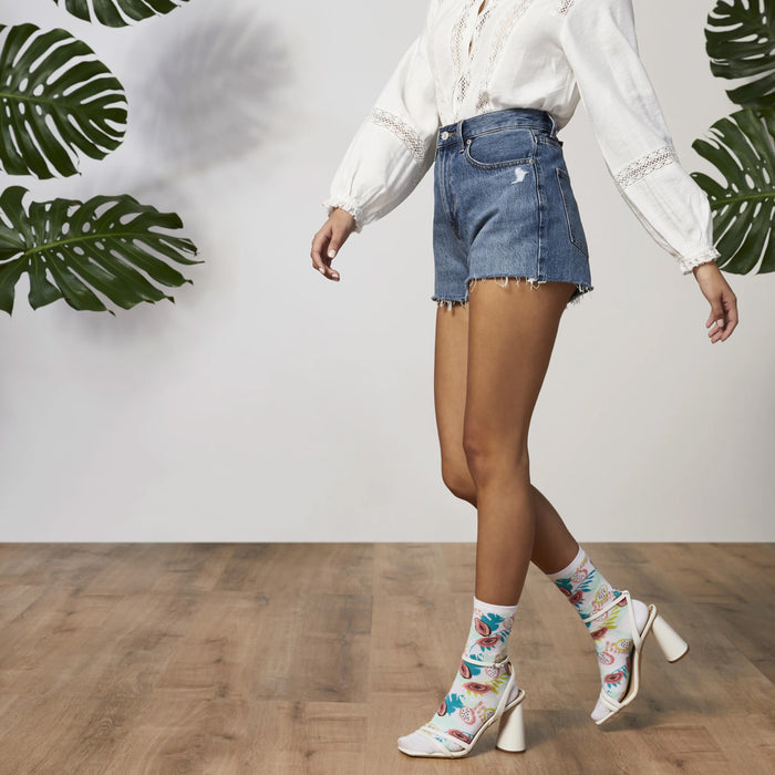 A model is shown wearing high-waisted denim shorts with a white blouse and white heeled sandals with tropical fruit printed socks.