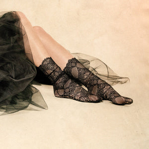 A pair of black lace socks displayed on a person's legs. The socks are mid-calf length and have a scalloped edge at the top. The person is wearing a black skirt made of a sheer material.