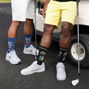 Lifestyle shot of two men on a golf course wearing the 'Swing Swear Repeat' socks and New Balance sneakers.