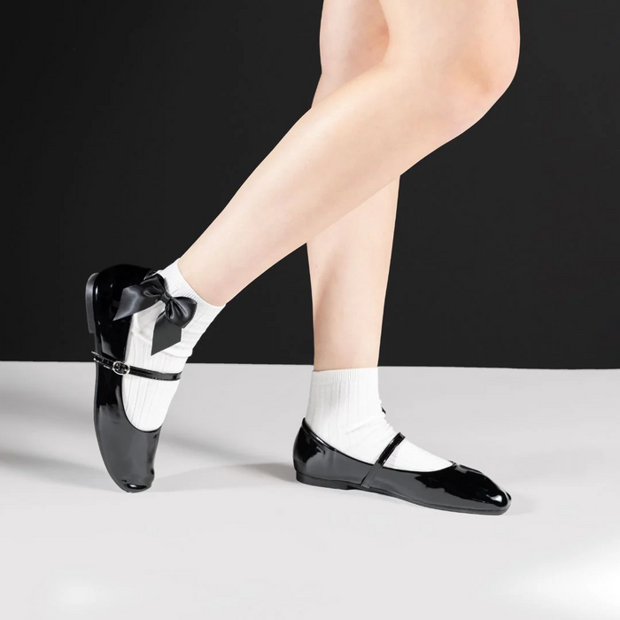 Lifestyle shot of a person wearing white ankle socks with a black bow and black patent leather shoes.