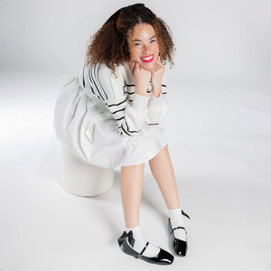 Overhead shot of a person wearing white ankle socks with a black bow and black patent leather shoes;  They're sitting down, smiling