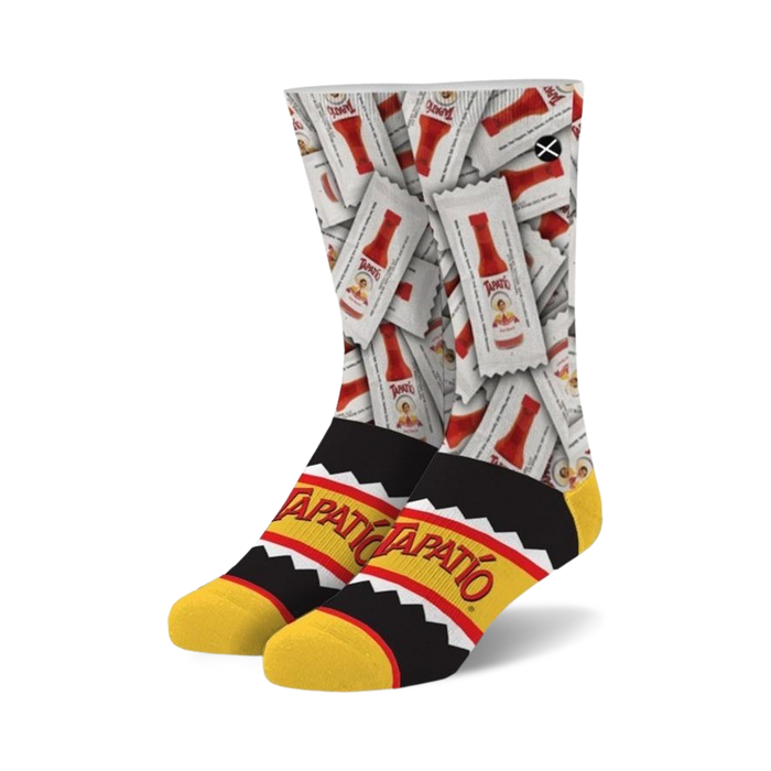 A pair of crew socks showcasing a repeating pattern of Tapatio hot sauce packets on a white background. The cuff features a black and yellow Tapatio logo.