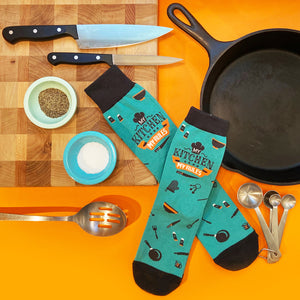Flatlay product shot: Teal 'My Kitchen My Rules' socks displayed with kitchen utensils and cutting board.