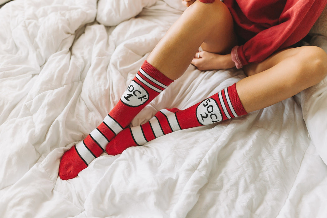 Lifestyle photo: A person wearing the red and white striped 'Sock 1 & 2' socks while sitting on a white bed.