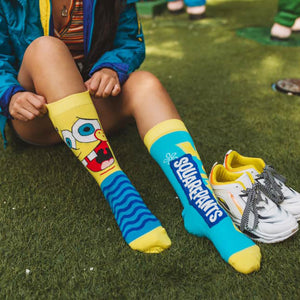 A person is shown from the knees down. They are putting on a pair of yellow socks that have a picture of Spongebob Squarepants on them. The socks also have blue and white striped cuffs. The person is wearing white sneakers with black and light blue laces.