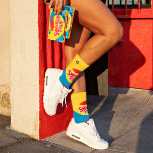 Lifestyle shot of a person's legs wearing the Swedish Fish socks and holding a box of the candy.  They're sitting against a red wall.