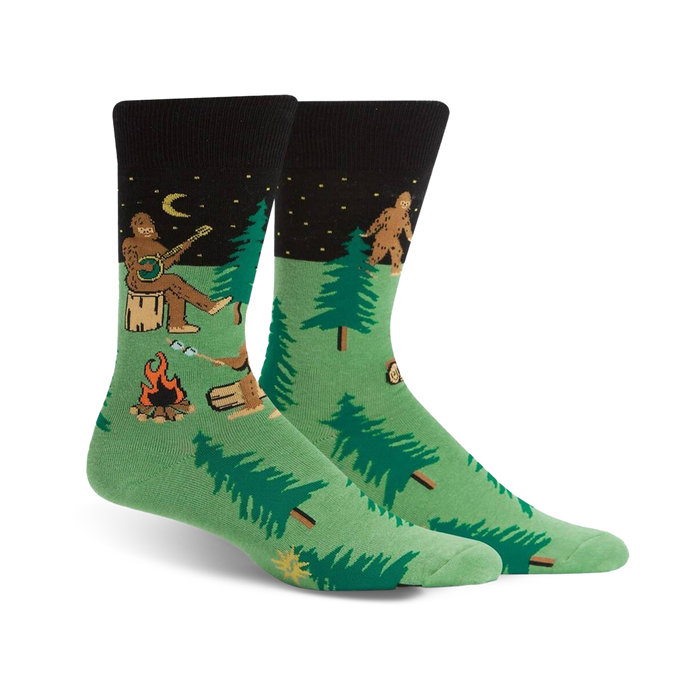 Pair of green and black crew socks featuring a whimsical Sasquatch playing banjo around a campfire in the woods.