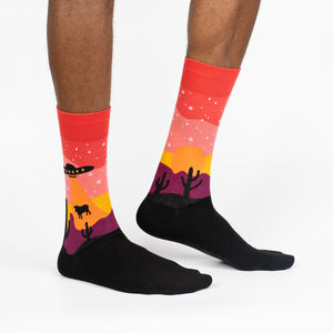 Lifestyle shot of a person wearing the Area 51 crew socks. Socks are red, orange, and purple with a desert scene featuring a UFO and cacti.