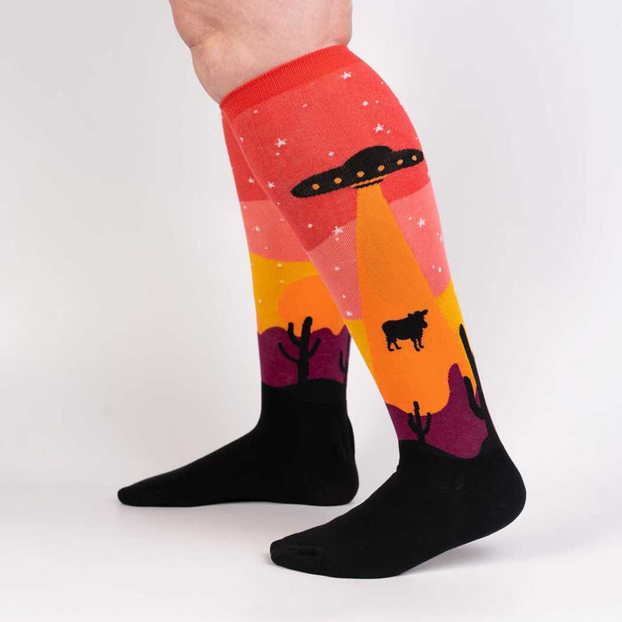 A pair of legs is shown from the back wearing a pair of knee-high socks with a Southwestern-inspired pattern including cacti, UFOs, and stars. The socks are red, orange, yellow, purple, and black.