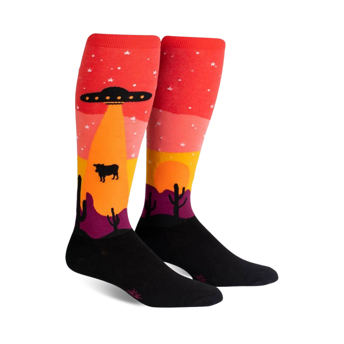 Pair of knee-high socks; red, orange, yellow, purple, and black. Design features an alien spaceship abducting a cow in a desert landscape at sunset.