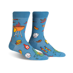 A pair of light blue crew socks with a fun BBQ design featuring burgers, hot dogs, grilling equipment, and chef hats.