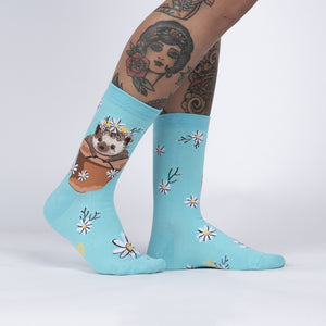 A pair of blue socks with a pattern of white daisies and green stems. On the left sock there is a cartoon illustration of a brown hedgehog wearing a flower crown sitting in a brown flower pot. The right sock does not have the hedgehog.