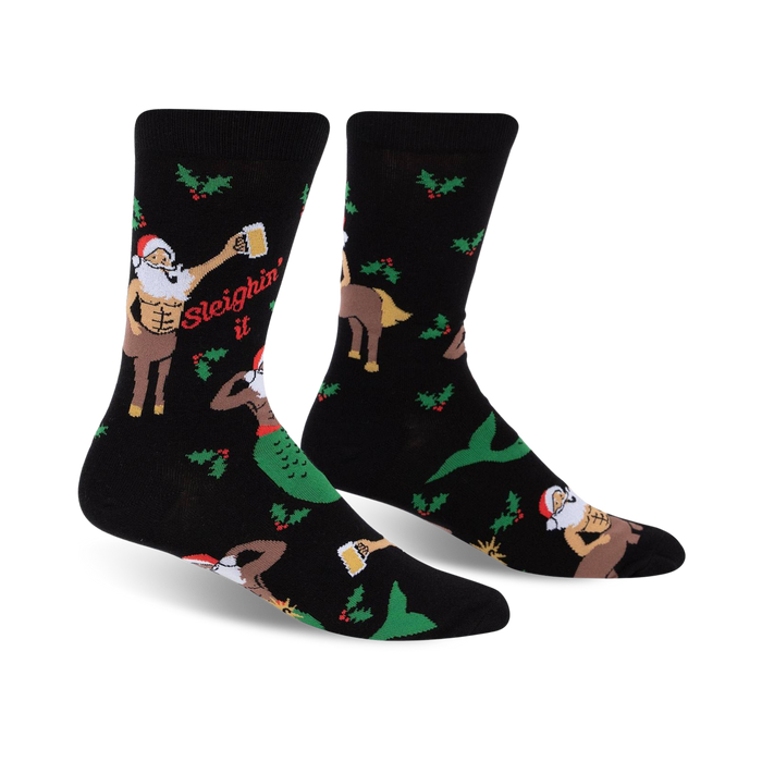 A pair of black crew socks with a festive design. The socks feature a muscular Santa centaur, a mermaid, holly, and the phrase 'Sleighin' It'. }}