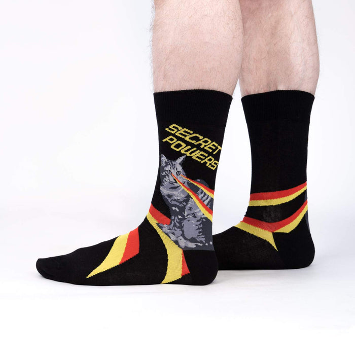 Lifestyle shot showing the 'Secret Powers' socks on a person's legs. The socks are predominantly black with yellow and red stripes.