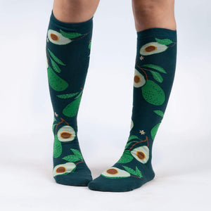 A pair of dark green knee-high socks with an all-over pattern of avocados, avocado leaves, and small white flowers.