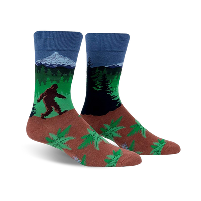 A pair of crew socks showcasing Bigfoot trekking through a forest landscape with a mountain in the background. The socks are predominantly blue, green, and brown, with a fern pattern at the bottom.