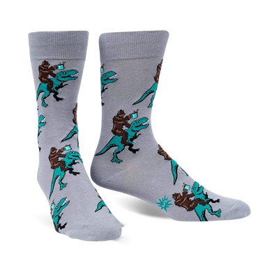 Pair of grey crew socks featuring a repeating pattern of Bigfoot riding a teal T-Rex and holding a mug.