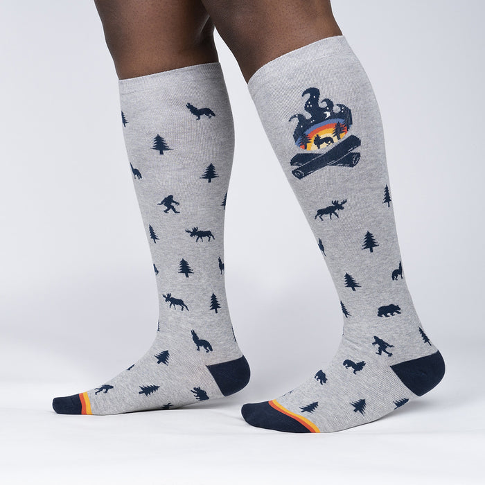 A pair of gray knee-high socks with a pattern of blue and orange pine trees, black silhouettes of bigfoot, and black and white silhouettes of moose and wolves. The socks have orange toes and heels and blue tops.