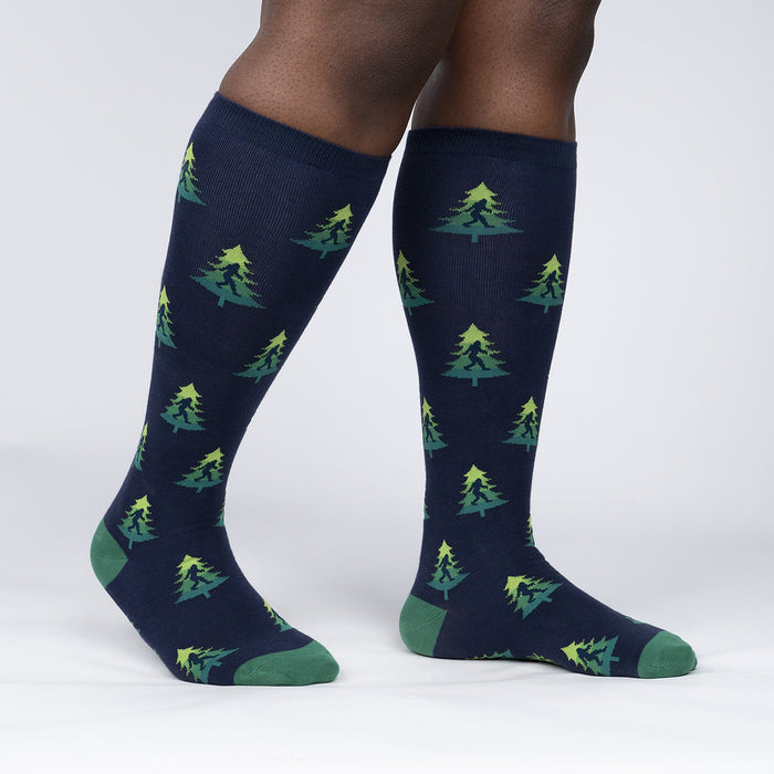 A pair of dark blue knee-high socks with a pattern of green pine trees and black silhouettes of Bigfoot.