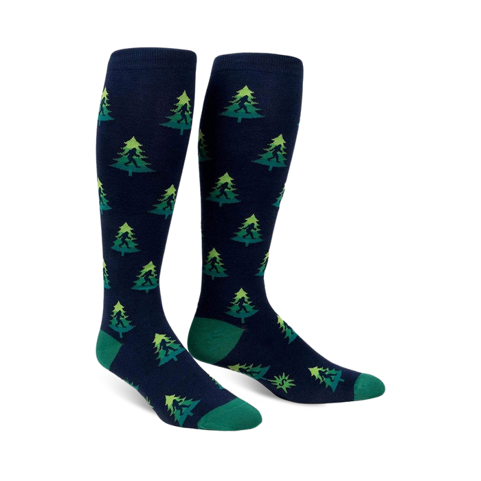 Pair of navy blue knee-high socks with a repeating pattern of green Bigfoot figures hidden amongst lime green and darker green Christmas trees.