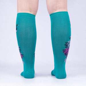 A pair of peacock-themed knee-high socks displayed on the back of a person's legs. The socks are teal with a multicolored peacock design on each leg.