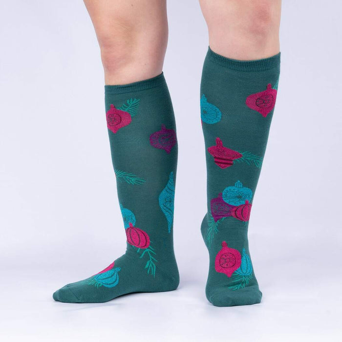 A pair of green knee-high socks with a pattern of pink and purple ornaments and pine branches.