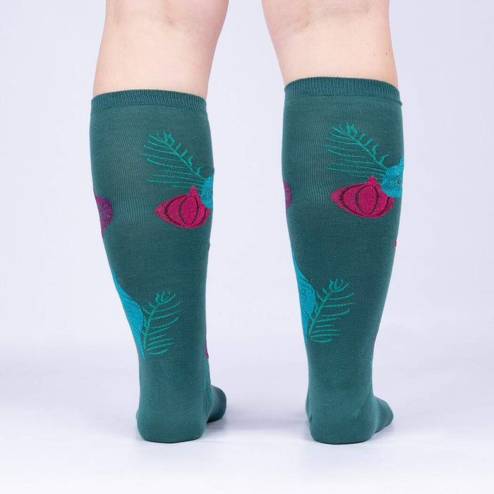 A pair of green knee-high socks with a pattern of pink and purple ornaments and pine branches.
