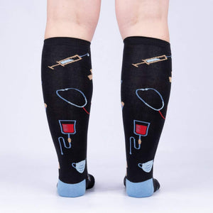 A pair of black knee-high socks with a pattern of medical instruments and supplies, including syringes, stethoscopes, IV bags, and masks.