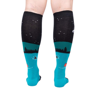 A pair of black knee-high socks with a blue toe and heel. The socks have a pattern of pine trees and a starry night sky with a red canoe on a lake.