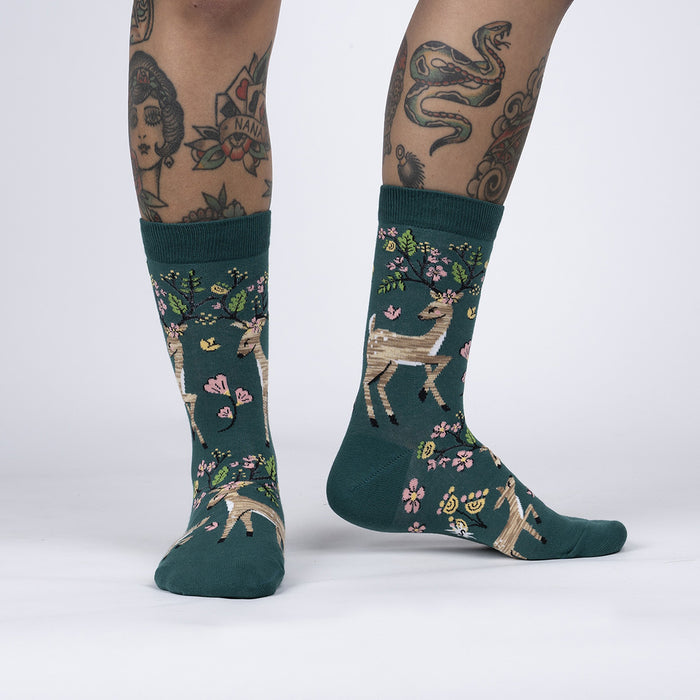 A pair of green knee-high socks with a pattern of deer, flowers, and vines on a white background. The socks are shown on a person's legs.